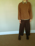 A person wearing a brown knit sweater with a black and white checkered collar, dark brown wide-leg trousers, and dark brown shoes, standing against a plain beige wall and carpeted floor.