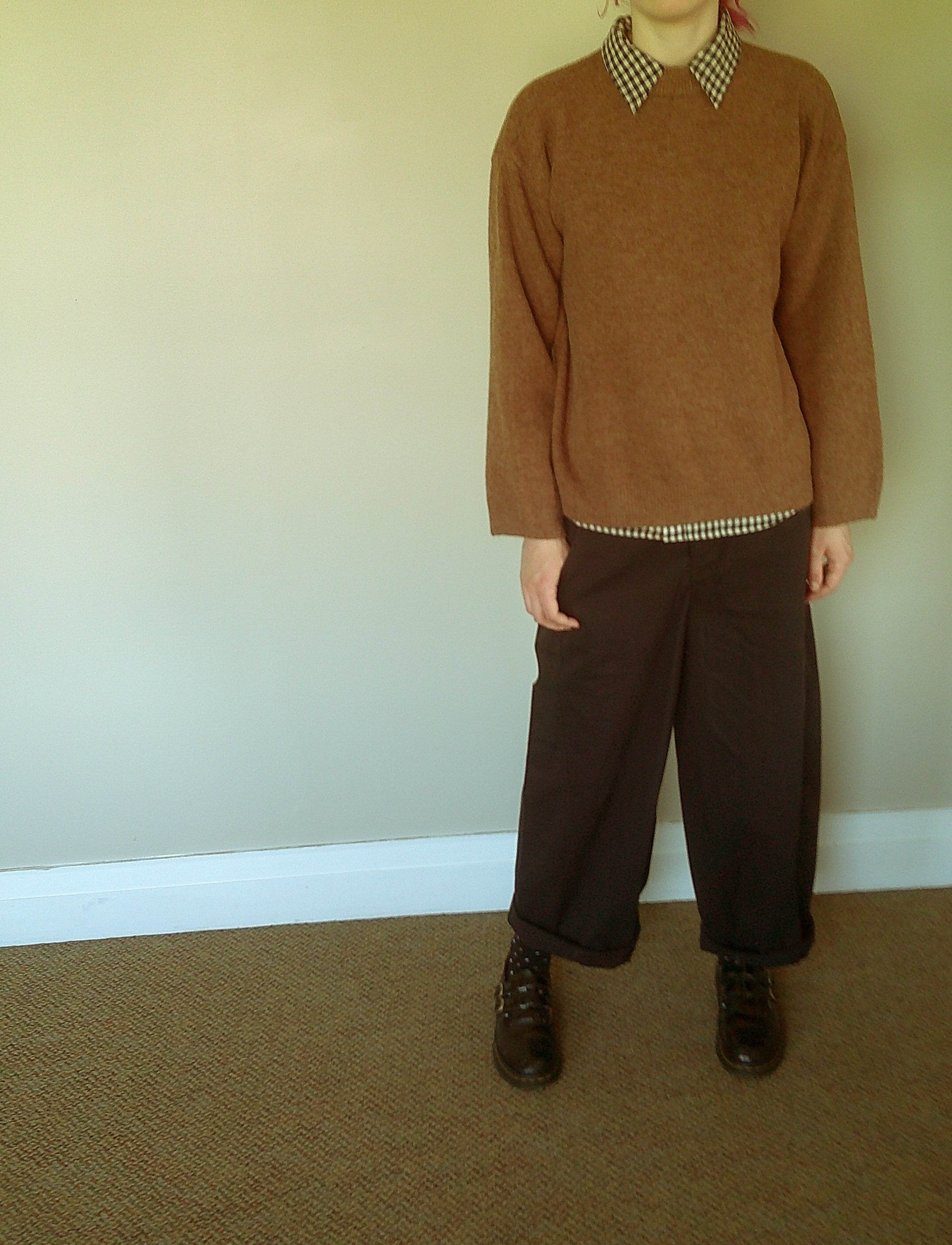 A person wearing a brown knit sweater with a black and white checkered collar, dark brown wide-leg trousers, and dark brown shoes, standing against a plain beige wall and carpeted floor.