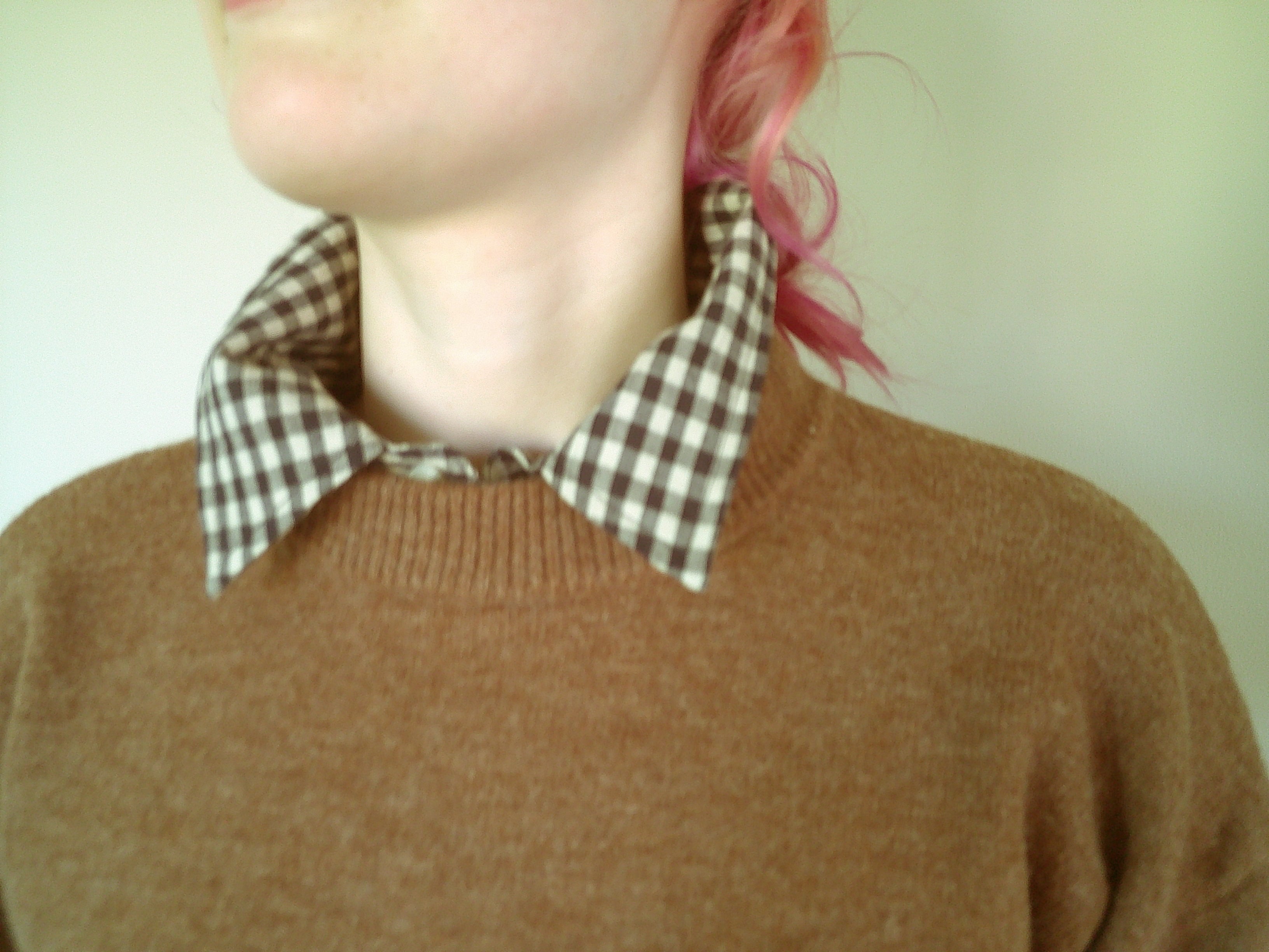 A close-up of a person wearing a brown knit crewneck sweater over a brown and white checkered collared shirt, with pink hair visible against a pale green background.