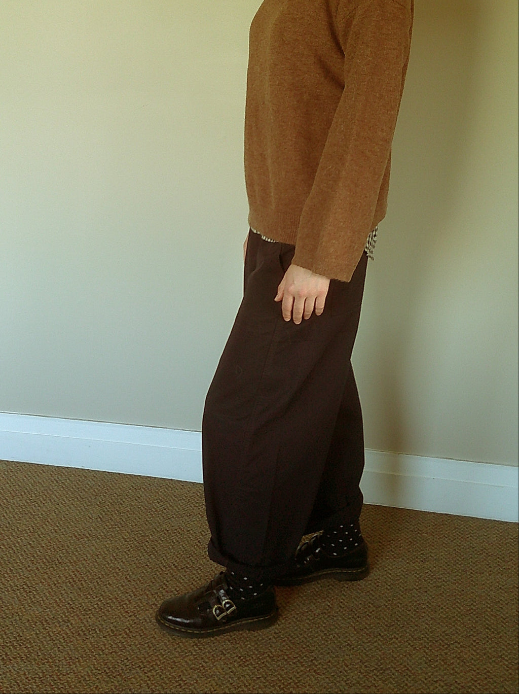 A person standing in profile wearing a brown knit sweater, dark brown wide-leg trousers, a checkered shirt collar, and dark brown shoes in an indoor room with a beige wall and carpeted floor.