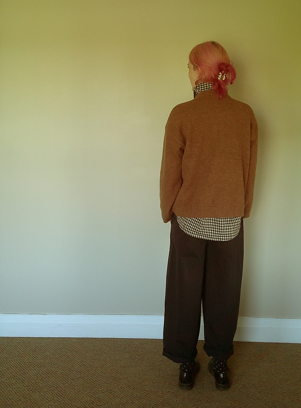 A person standing with their back to the camera, wearing a brown knit sweater over a black and white gingham shirt, loose dark brown trousers, and black shoes, set against a plain beige wall.