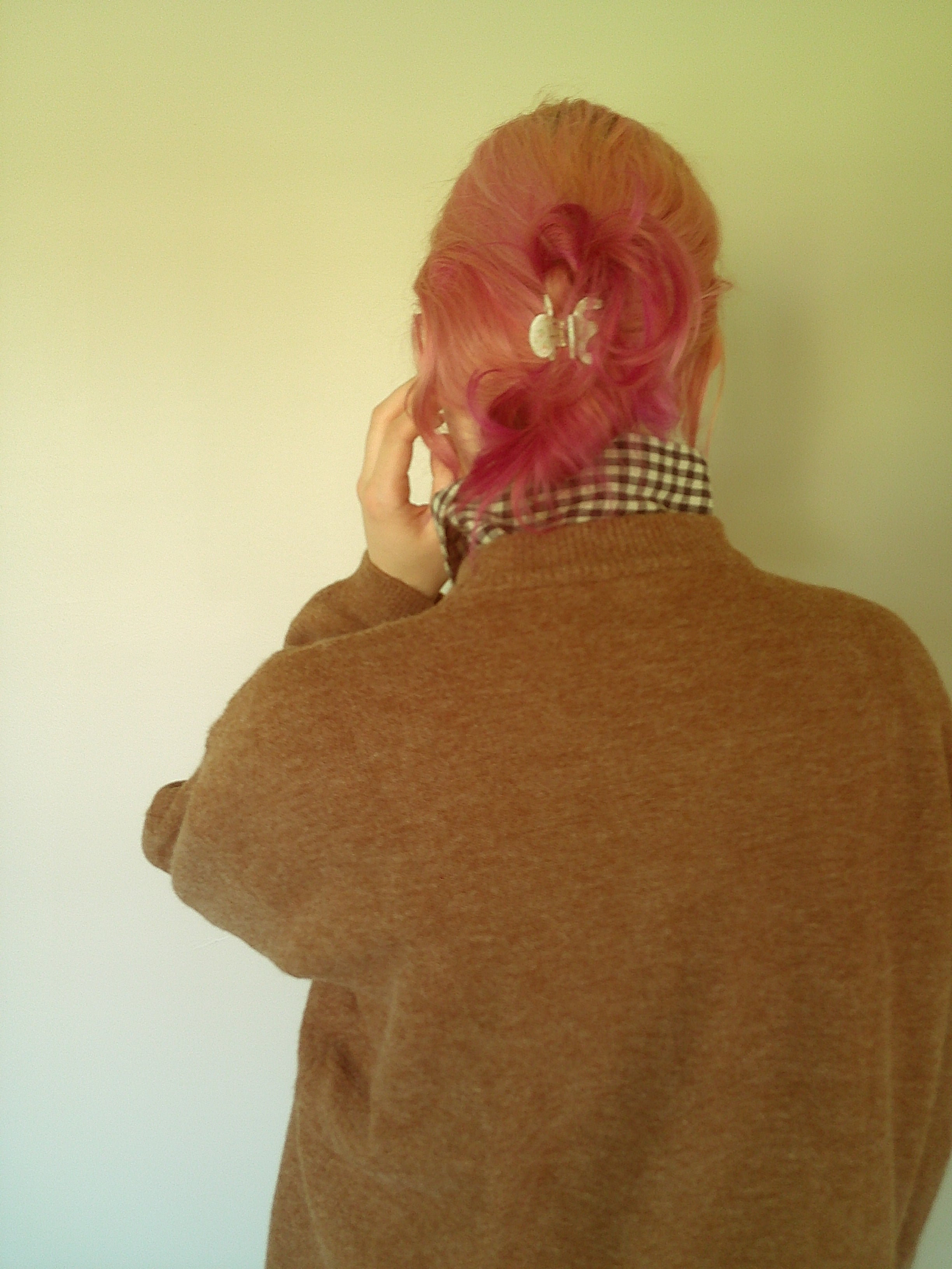 A person shown from behind wearing a brown knit sweater, a black and white checkered shirt, and bright pink hair, set against a plain light yellow wall.