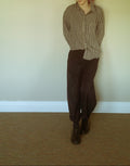A person standing in a room wearing a brown and beige gingham button-up shirt, dark brown pants, and dark brown lace-up boots, against a plain beige wall and carpeted floor.