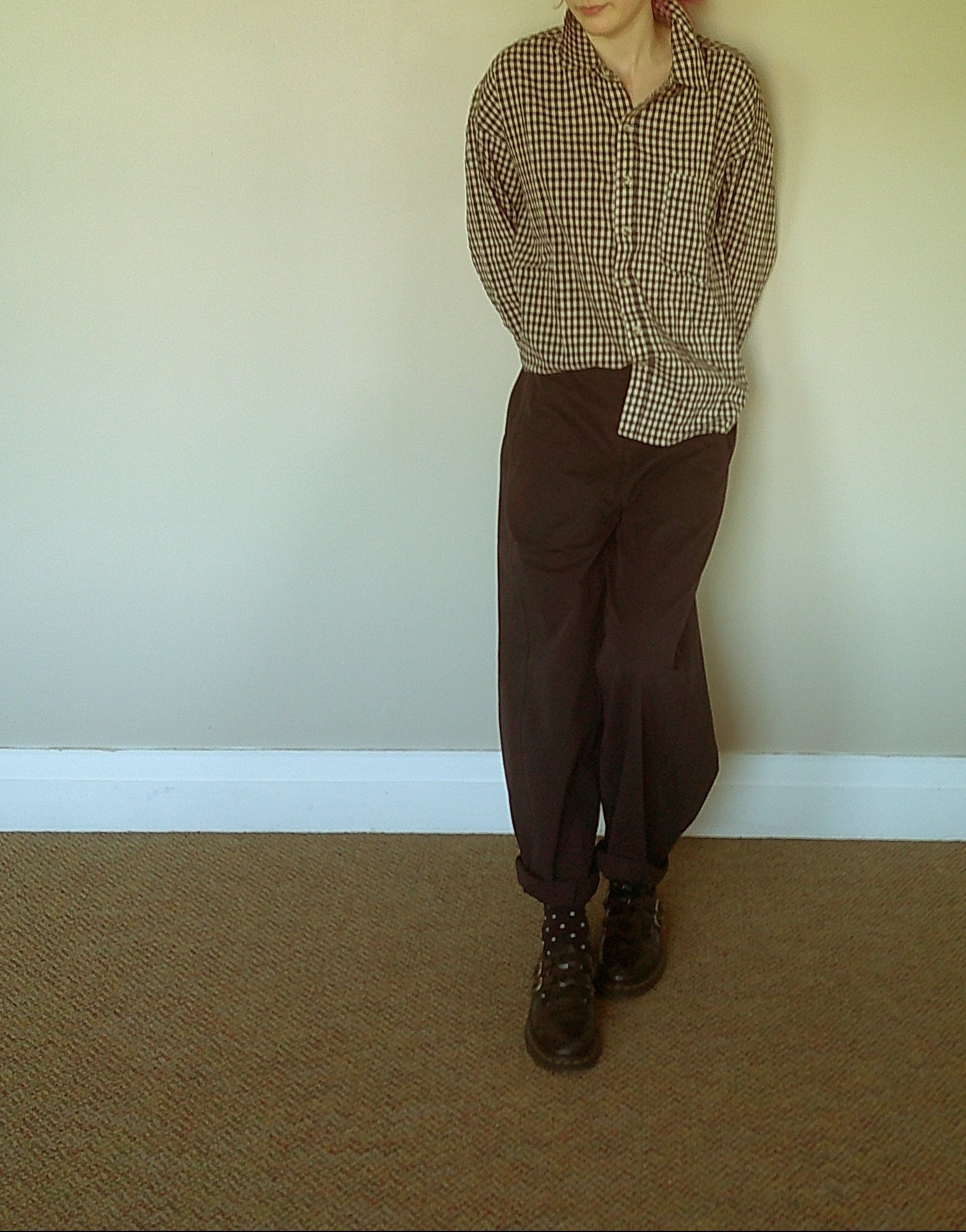 A person standing in a room wearing a brown and beige gingham button-up shirt, dark brown pants, and dark brown lace-up boots, against a plain beige wall and carpeted floor.