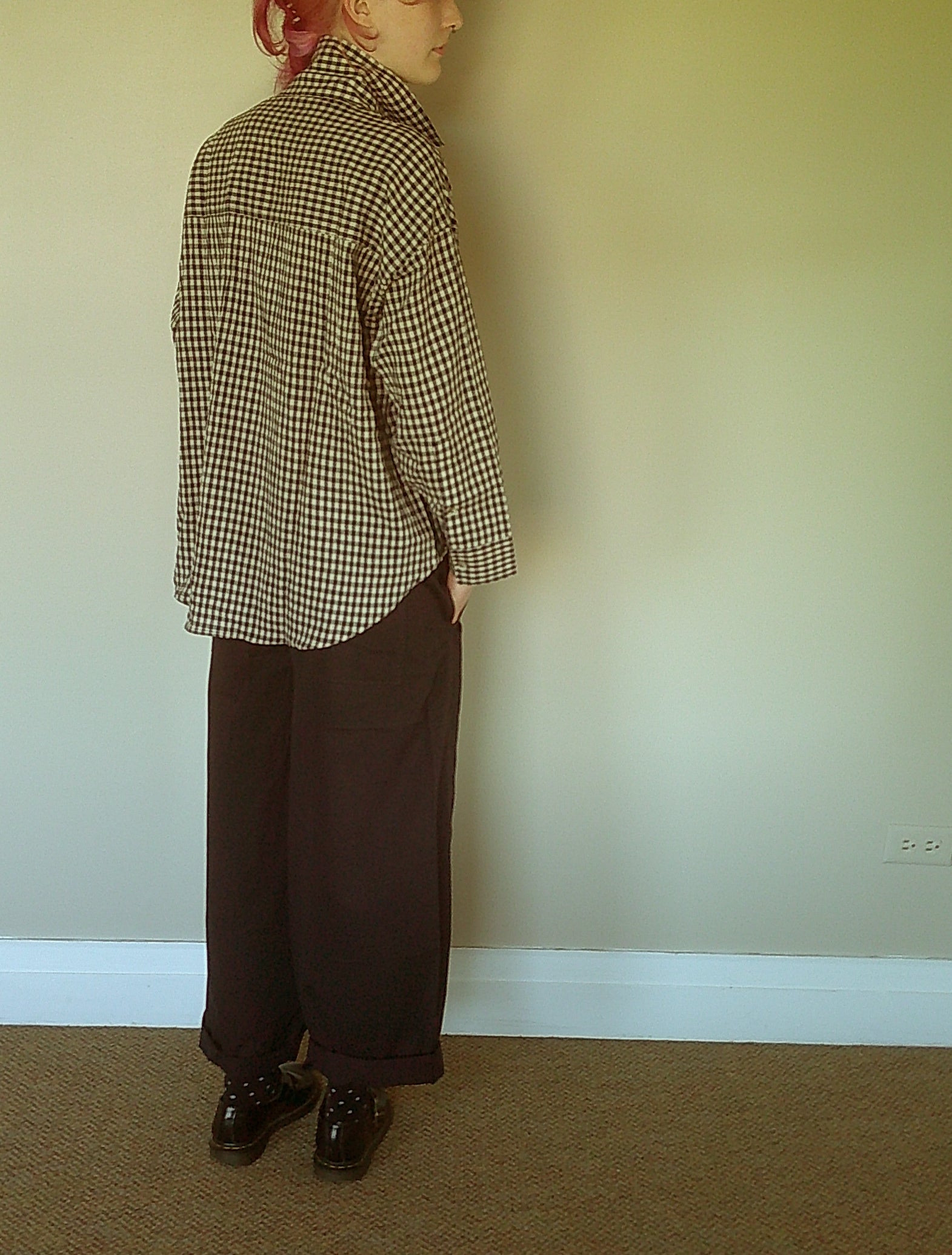 A person standing in profile wearing a blue and beige gingham long-sleeve shirt, dark brown wide-leg trousers, and dark brown shoes, set against a plain beige wall and carpeted floor.