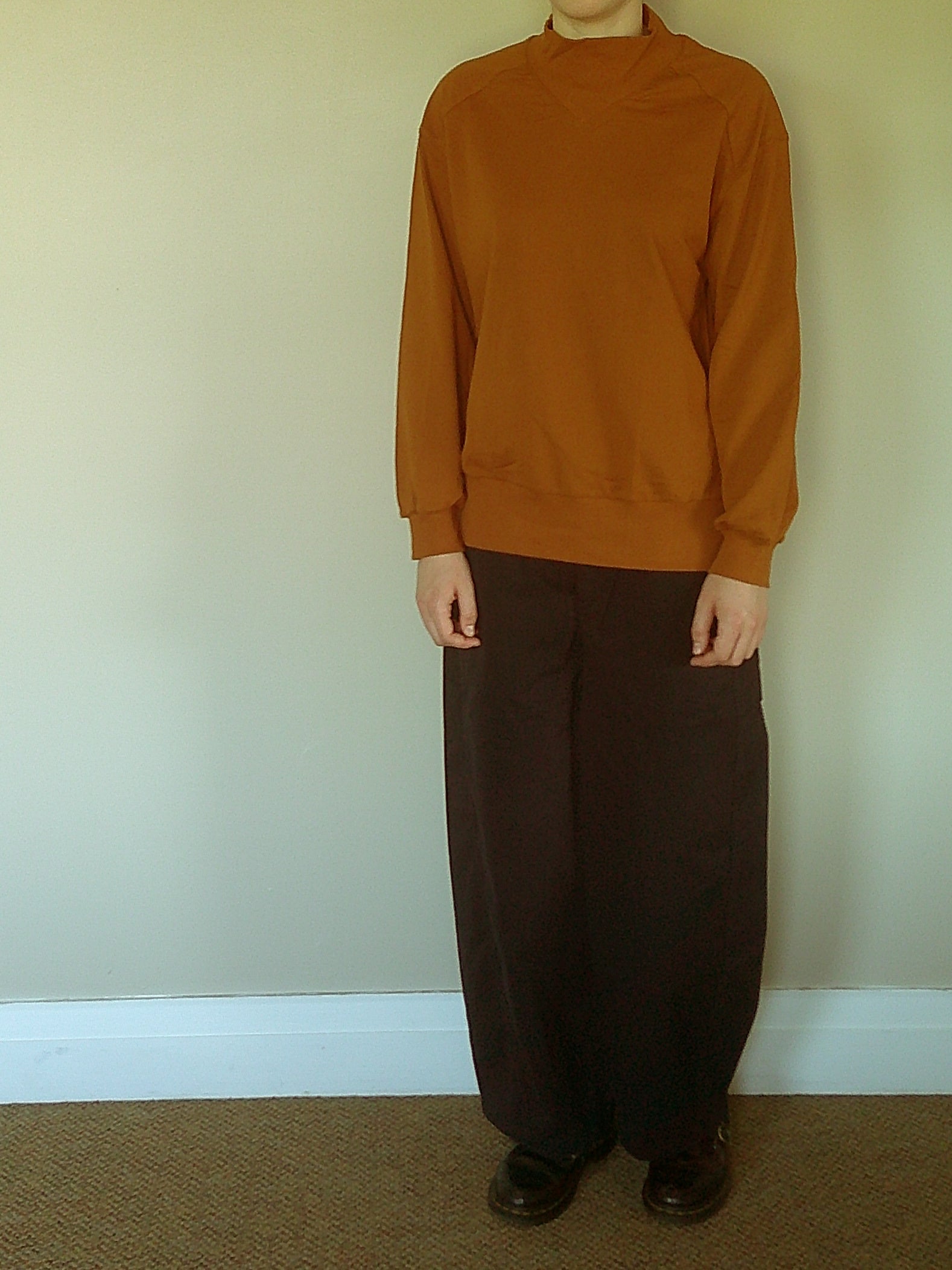 A person standing indoors wearing a rust-orange long-sleeve mock-neck shirt and dark blue wide-leg pants with black shoes, against a plain beige wall.
