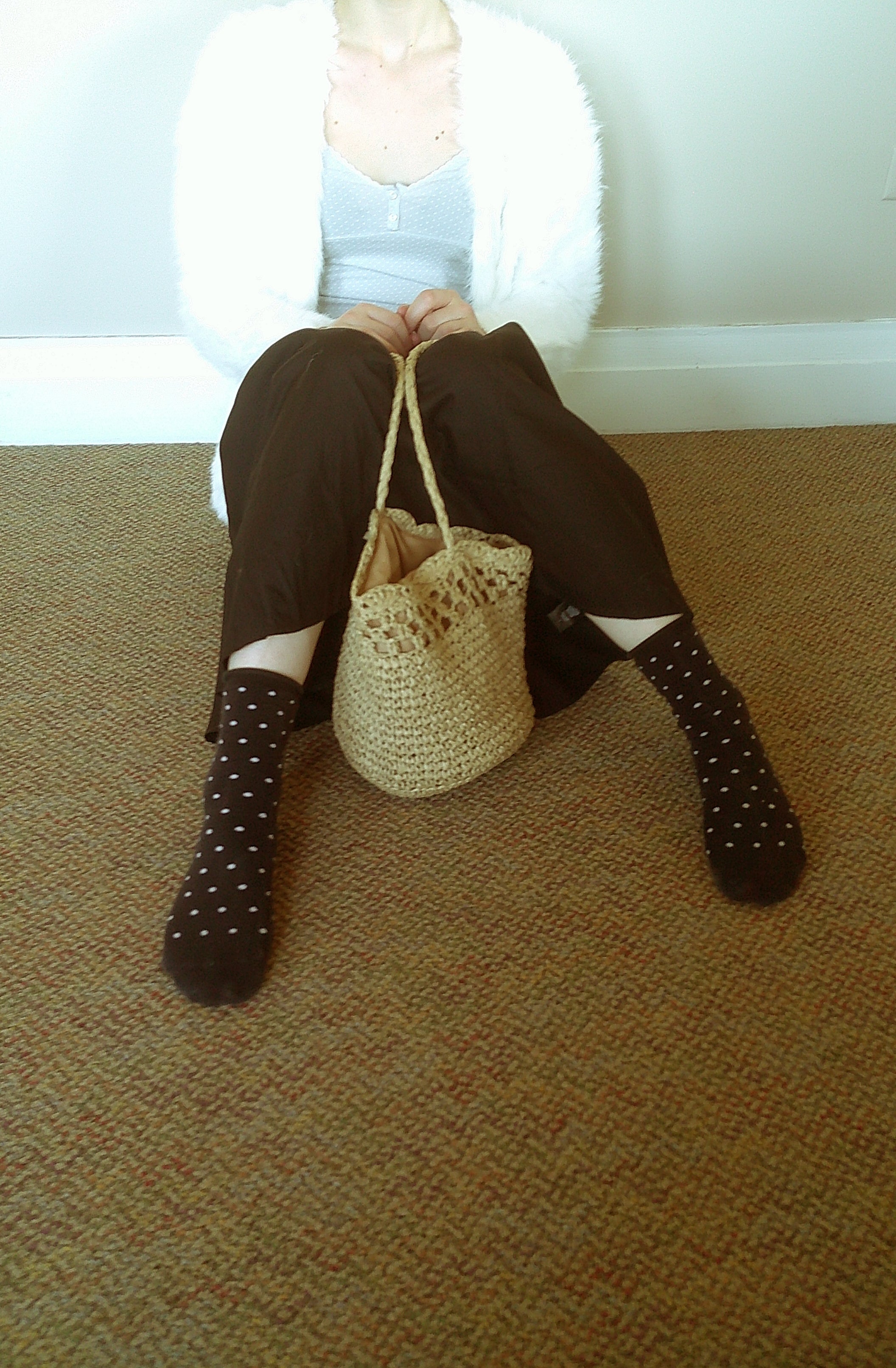 A person sitting on a carpeted floor wearing dark brown pants, black socks with white polka dots, a white cardigan, and a white tank top, holding a small woven straw tote bag.