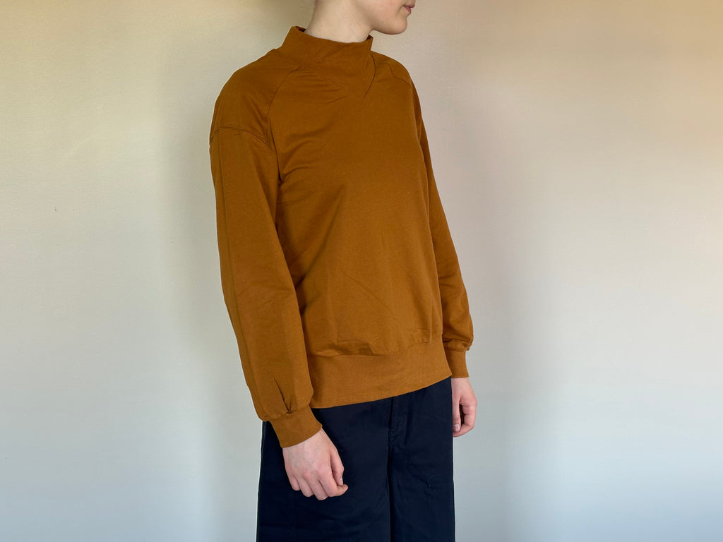 A person shown in side profile wearing a long-sleeve mock-neck knit top in a dark mustard color, paired with dark pants, against a plain light wall.