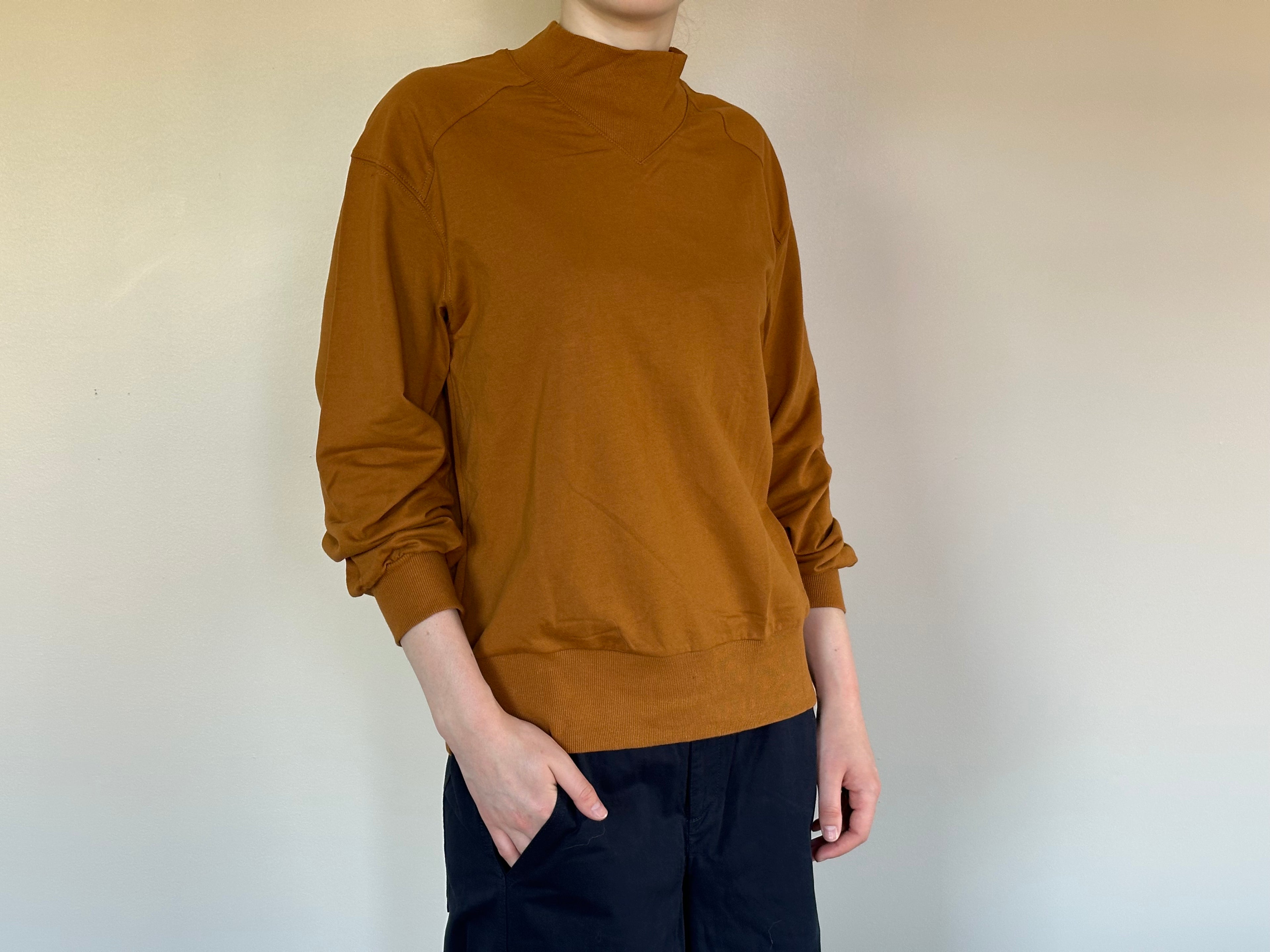 A person wearing a long-sleeve mock neck knit sweater in a dark mustard color, paired with dark pants, standing against a plain off-white wall.