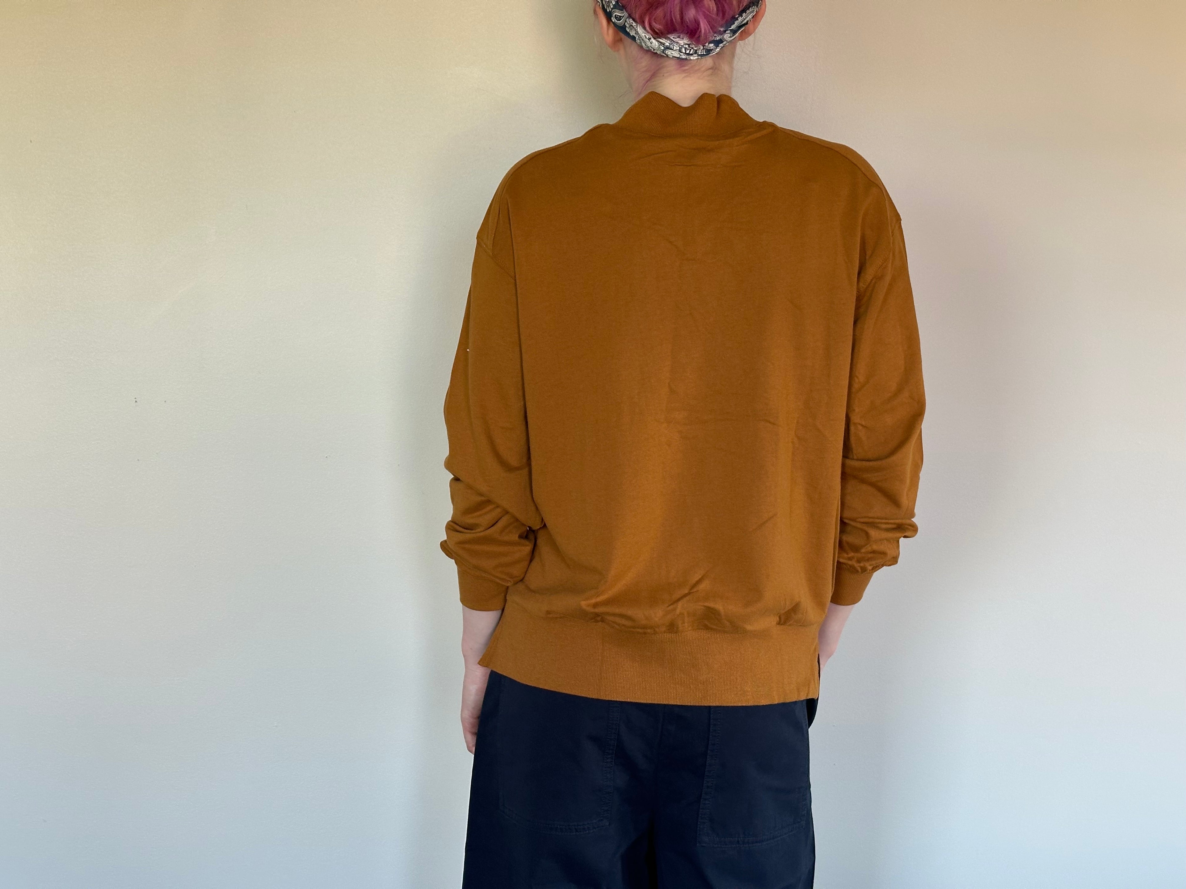 A person shown from the back wearing a mustard brown mock-neck long-sleeve top and dark pants, standing against a plain off-white wall.