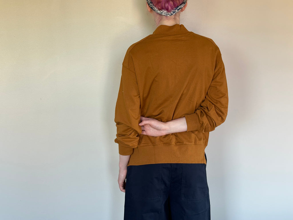 A person shown from the back wearing a mustard brown long-sleeve mock-neck top and dark pants, standing against a plain off-white wall.