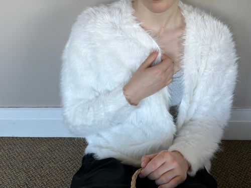 A person wearing a fluffy white faux-fur cardigan or jacket over a gray top and black pants, seated indoors against a plain wall and carpeted floor.