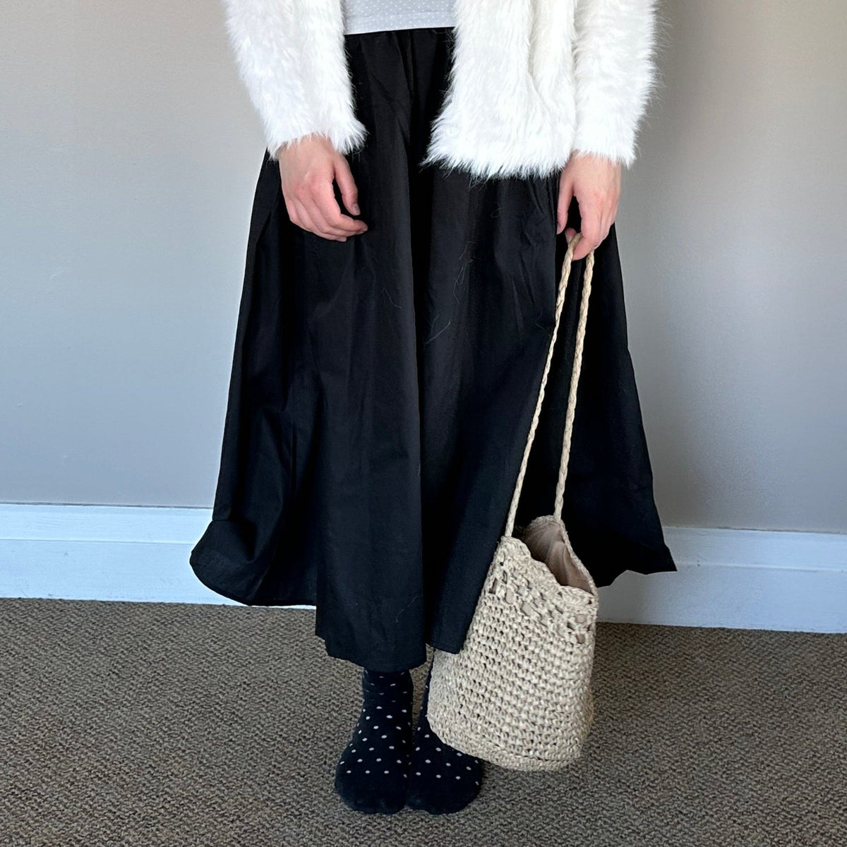 A person standing indoors wearing a fluffy white cardigan over a light gray tank top, a long black skirt, black polka-dot socks, and holding a woven beige tote bag against a plain gray wall.