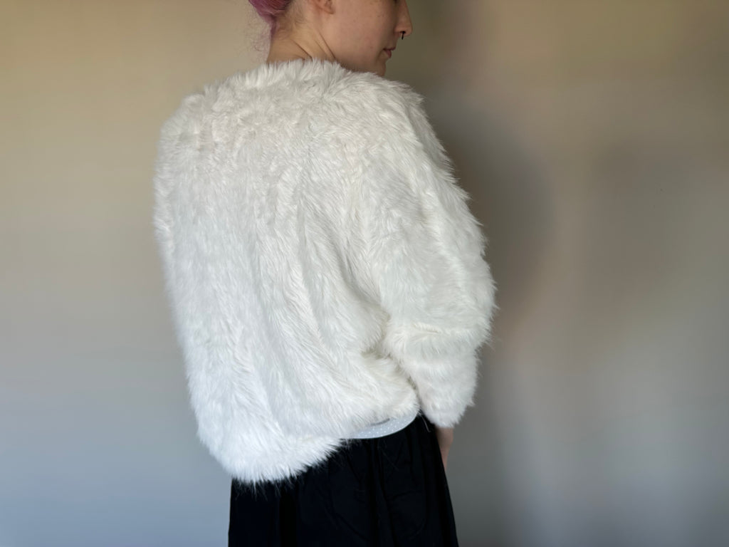 A person shown from the back wearing a fluffy white sweater over a black skirt, standing against a plain indoor wall.