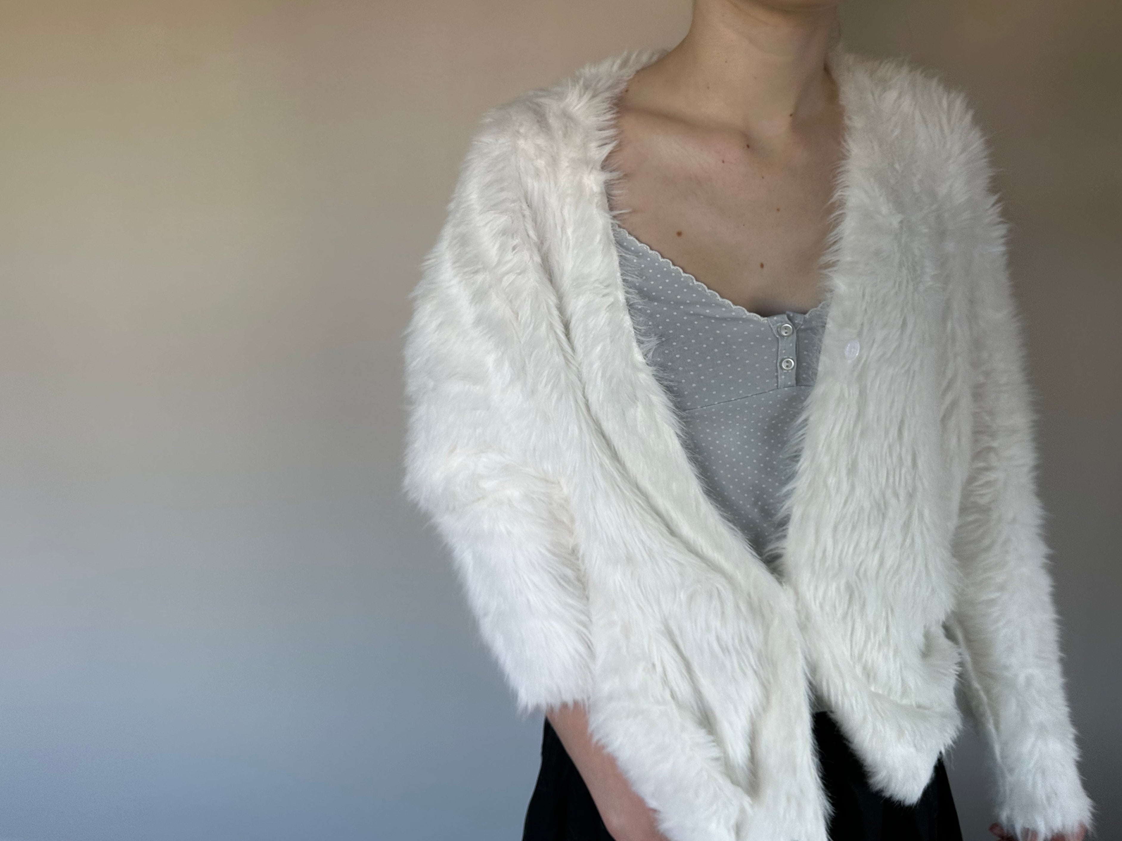 A person wearing a fluffy white faux-fur cardigan over a gray knit top and black bottoms, standing against a plain light gray wall.
