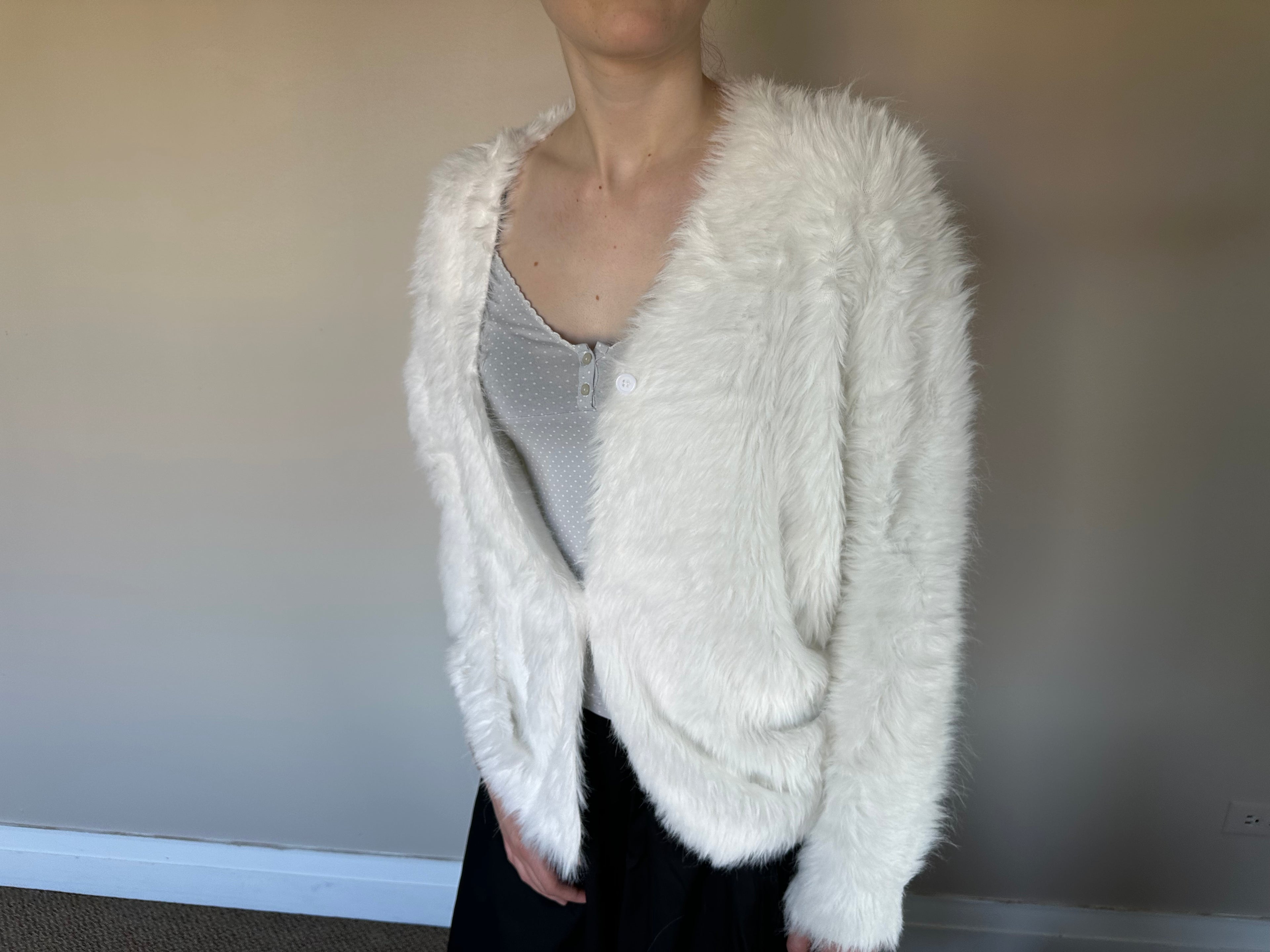 A person wearing a fluffy white faux-fur cardigan over a light gray top and black bottoms, standing against a plain indoor wall.