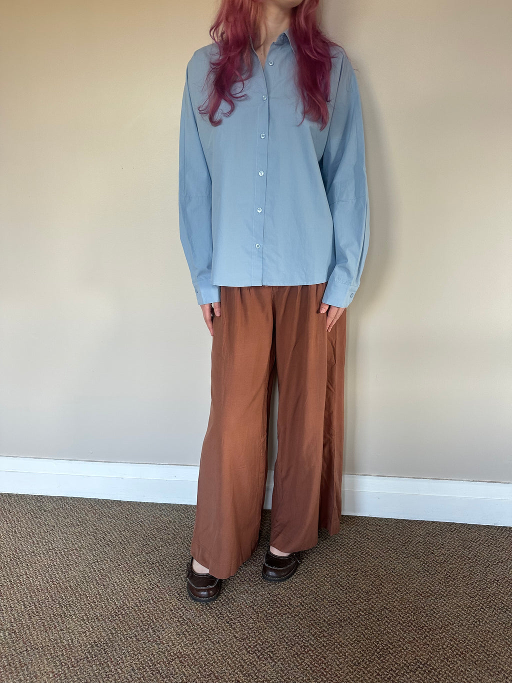 A person wearing a light blue long-sleeve button-up shirt and wide-leg rust-brown pants, standing against a plain beige wall with carpeted floor.