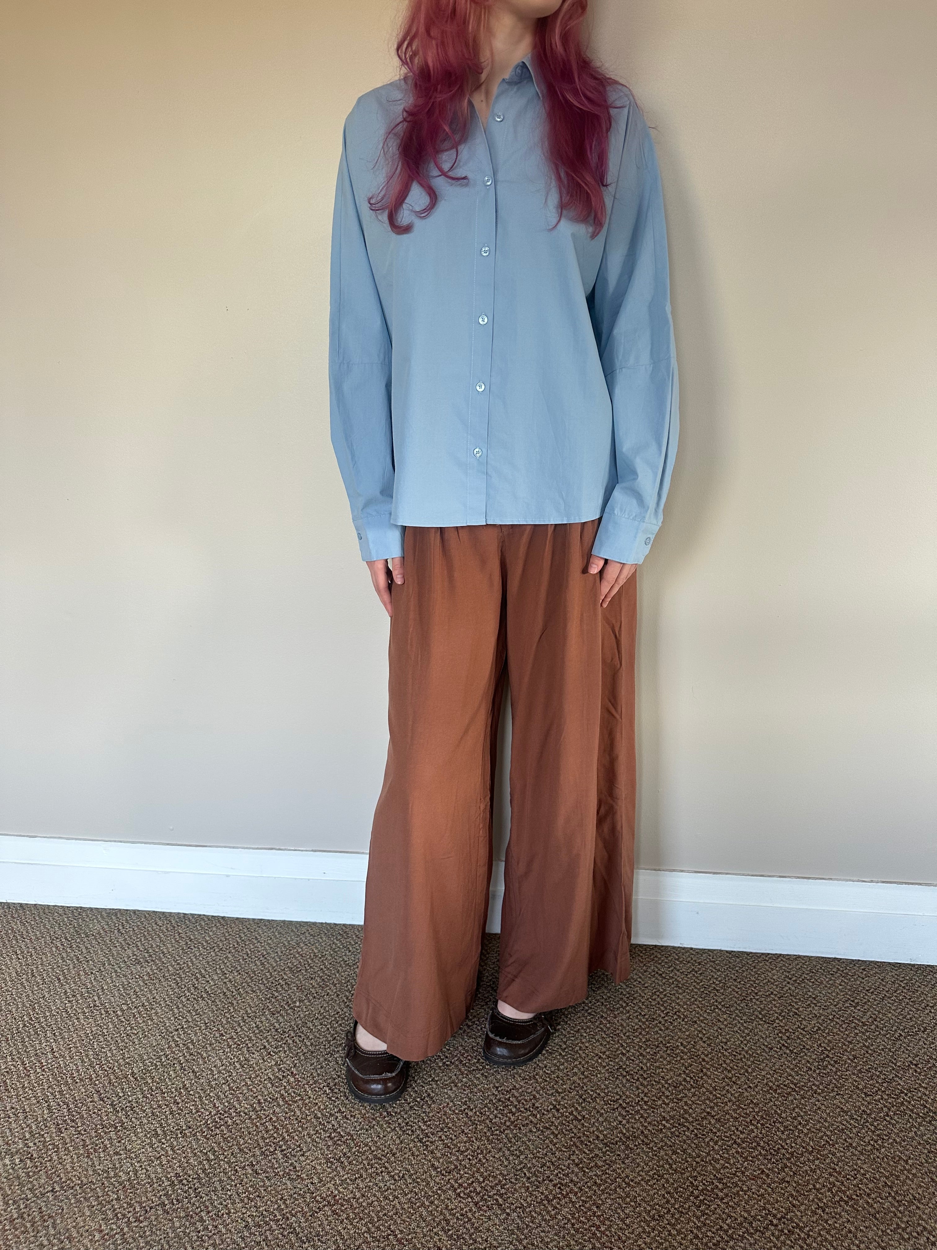 A person wearing a light blue long-sleeve button-up shirt and wide-leg brown pants, standing against a plain beige wall with carpeted floor.