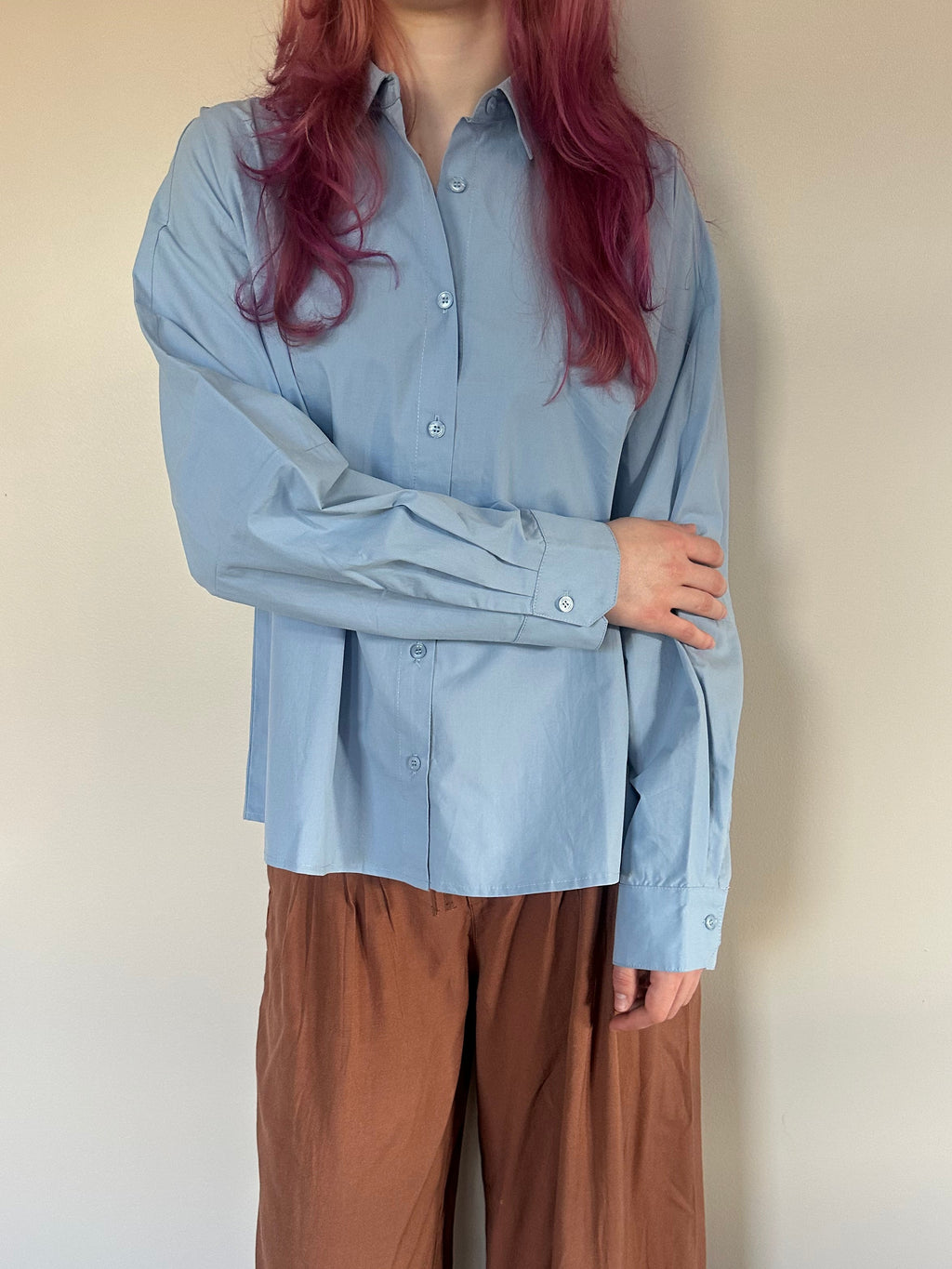 A person wearing a light blue long-sleeve button-up shirt and brown wide-leg pants, standing against a plain beige wall.