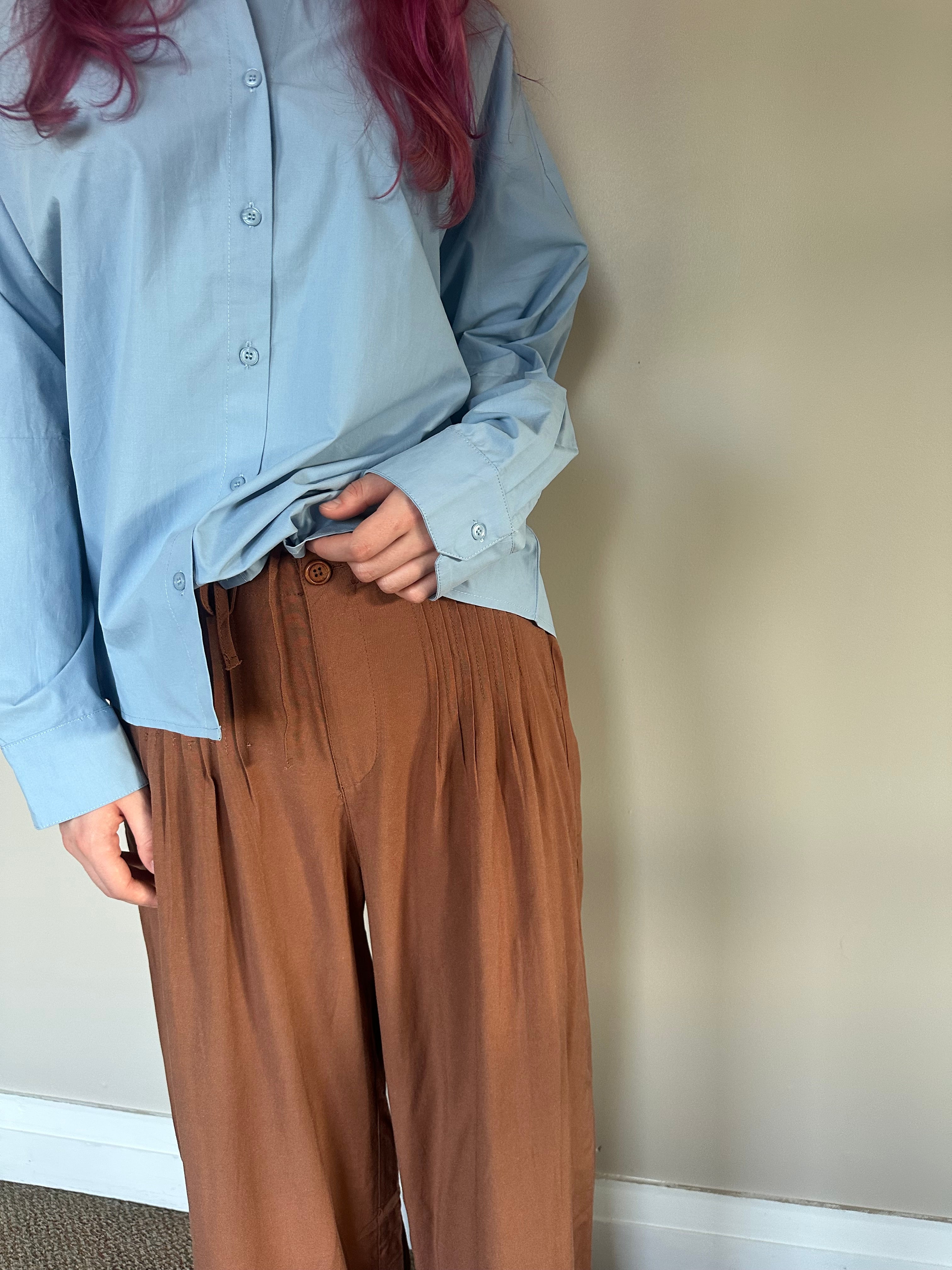A person wearing a light blue long-sleeve button-up shirt and brown wide-leg pleated trousers, standing against a plain beige wall.