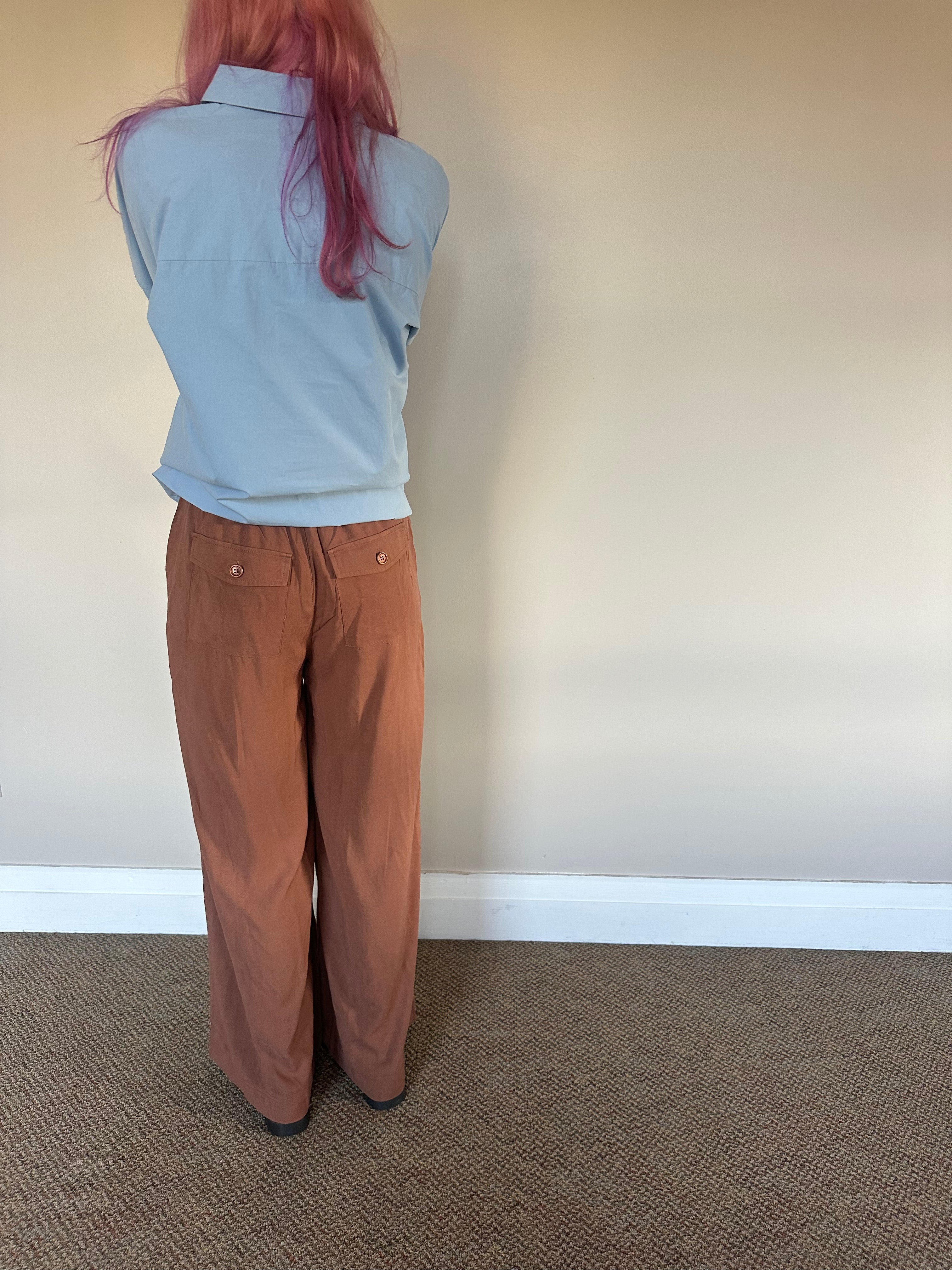 A person standing with their back to the camera, wearing a light blue sleeveless collared top and rust-brown wide-leg pants, in front of a plain beige wall and carpeted floor.