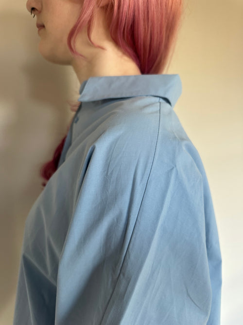 A close-up side profile of a person with pink hair wearing an oversized light blue button-up shirt against a plain beige wall.