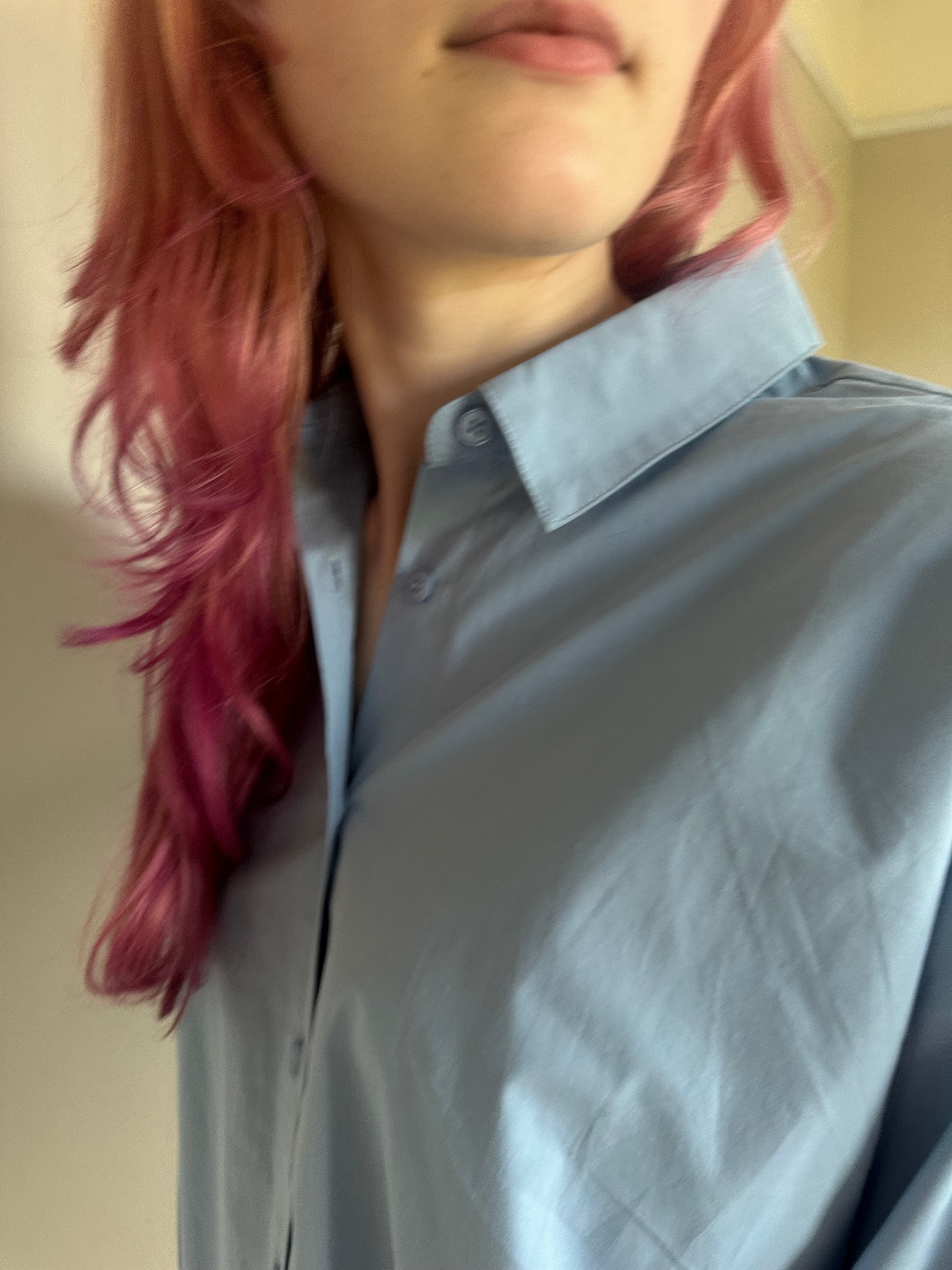 A close-up of a person wearing a light blue button-down shirt, showing the collar, front placket, and shoulder area. The person has pink hair, and the background is a plain indoor wall.