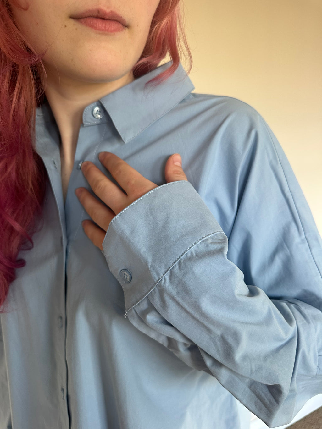 A close-up of a person wearing a light blue long-sleeve button-up shirt, with their hand resting on the chest. The person has pink hair, and the background is a plain beige wall.