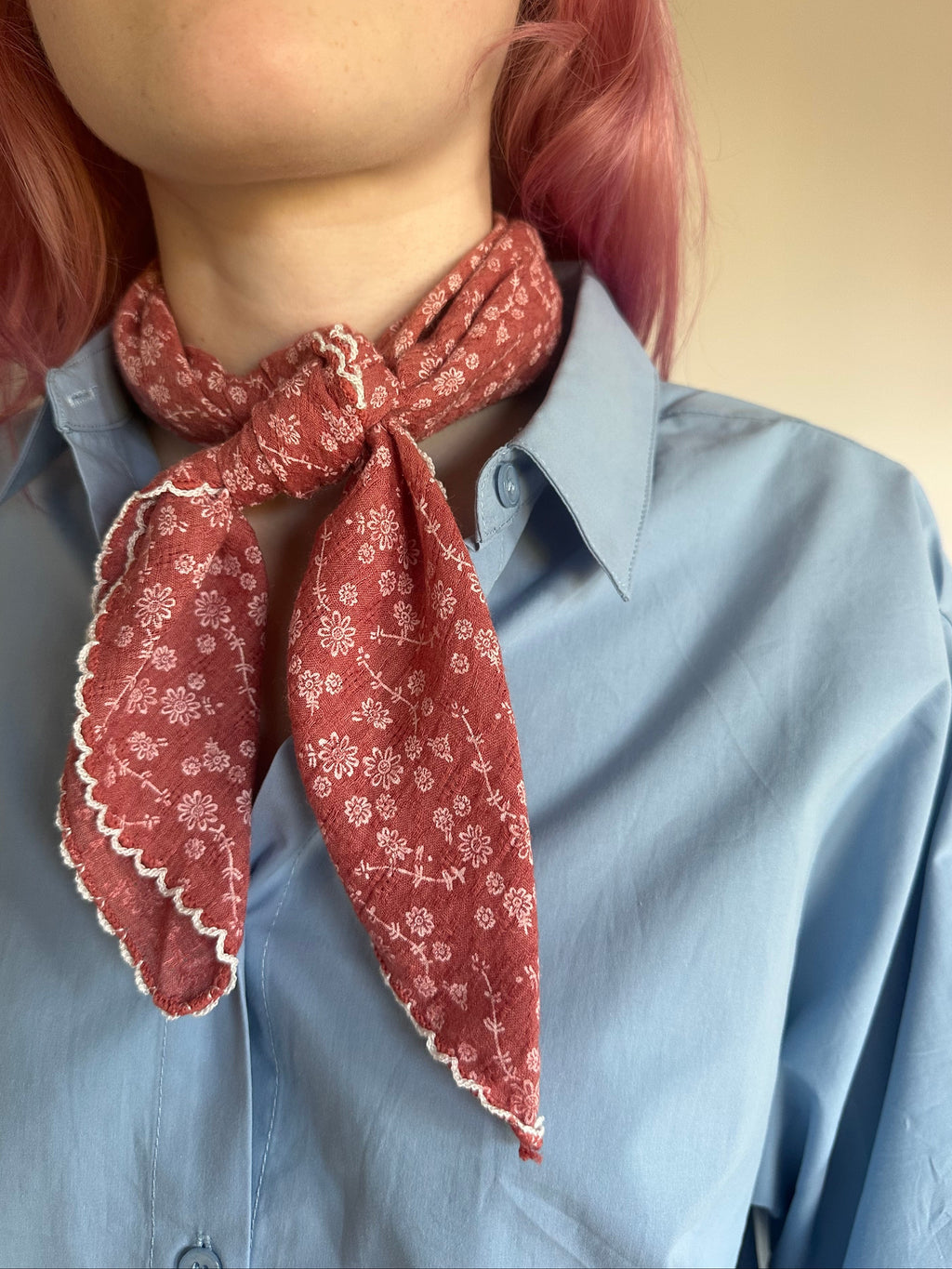 A close-up of a person wearing a dusty red patterned neck scarf tied around a light blue button-down shirt. The scarf features a delicate white floral print and scalloped edges.