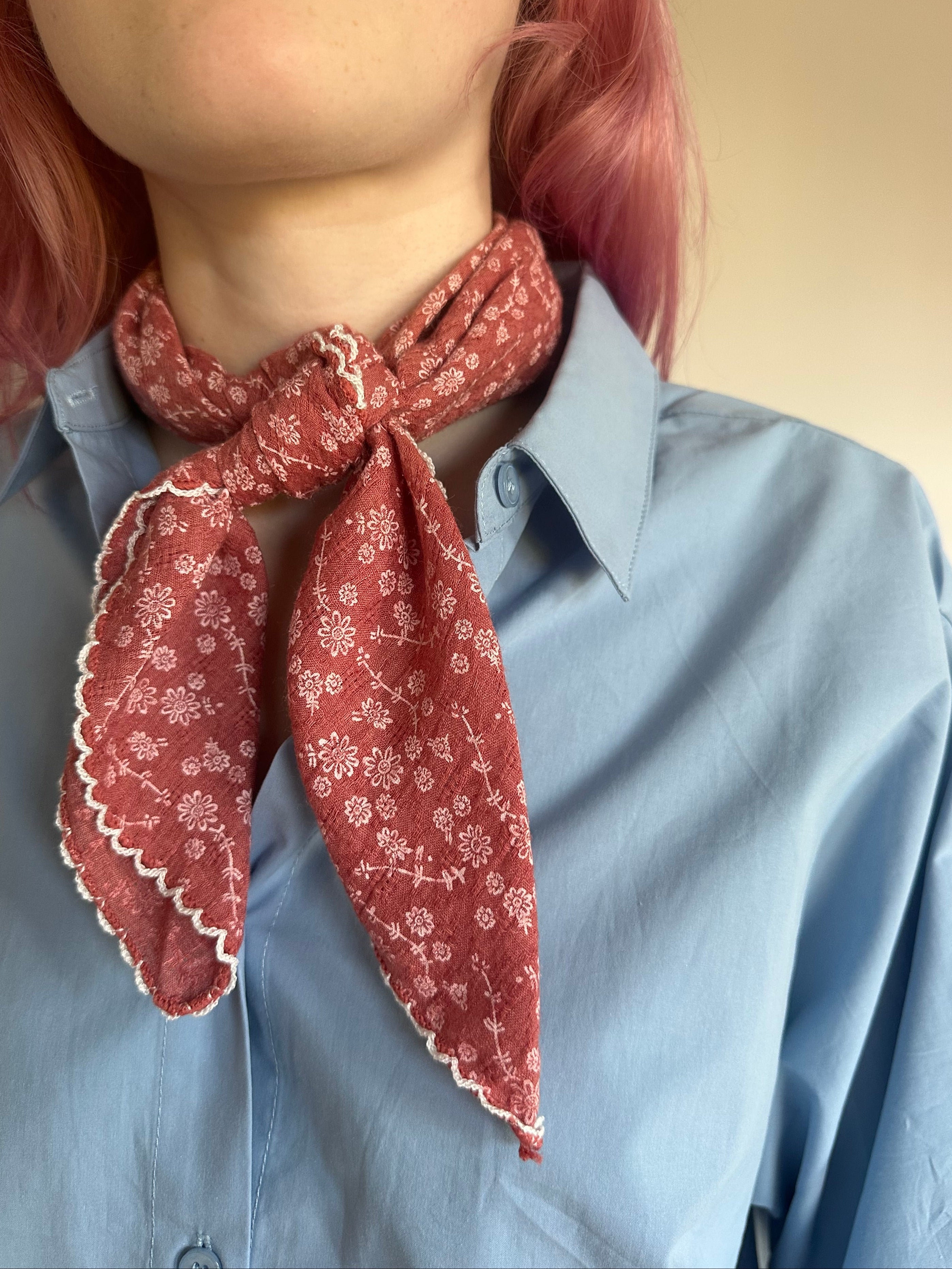 A close-up of a person wearing a dusty red patterned neck scarf tied around a light blue button-down shirt. The scarf features a delicate white floral print and scalloped edges.