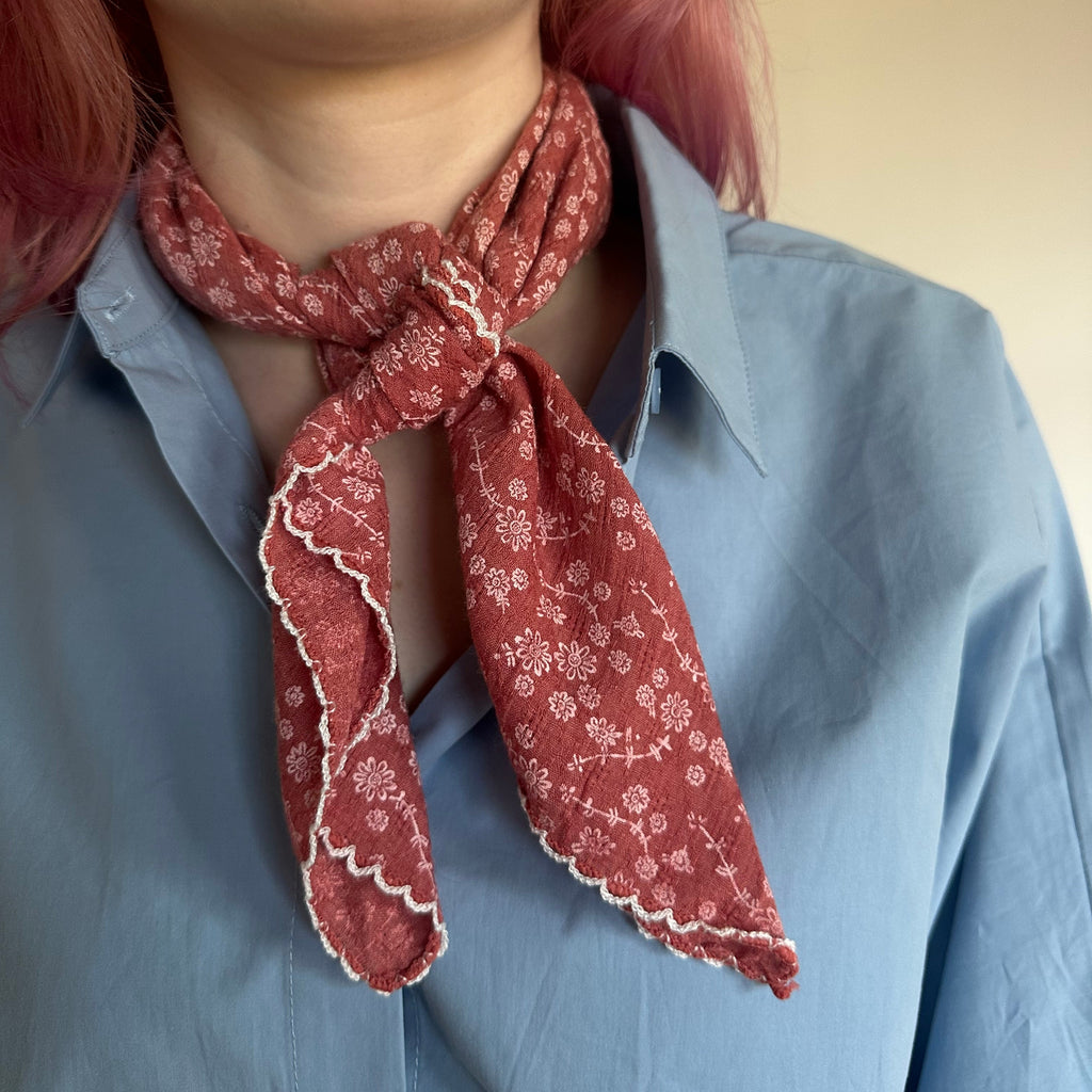 A close-up of a person wearing a dusty red patterned neck scarf tied around a light blue button-down shirt. The scarf features a white floral print and scalloped edges.