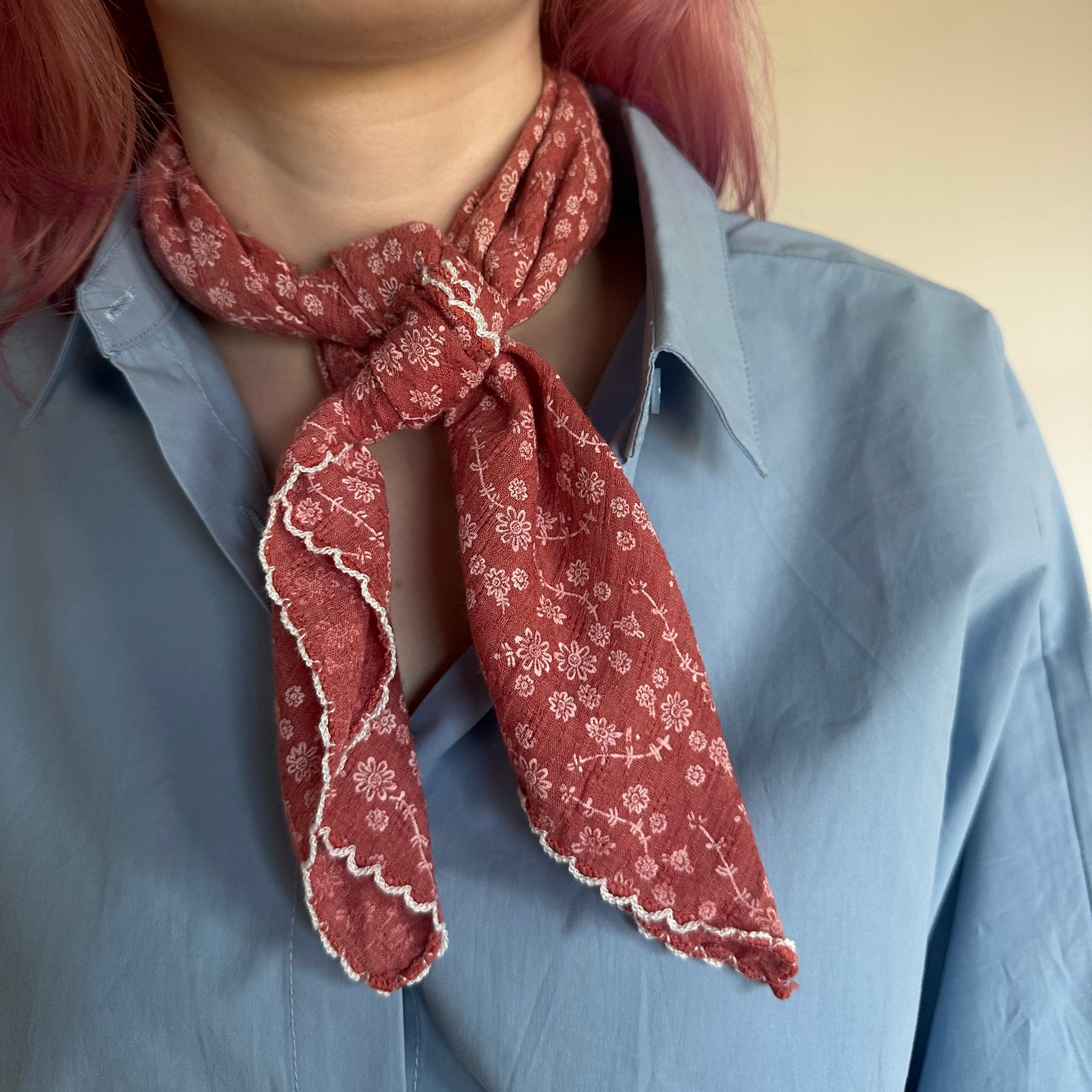 A close-up of a person wearing a dusty red patterned neck scarf tied around a light blue button-down shirt. The scarf features a white floral print and scalloped edges.