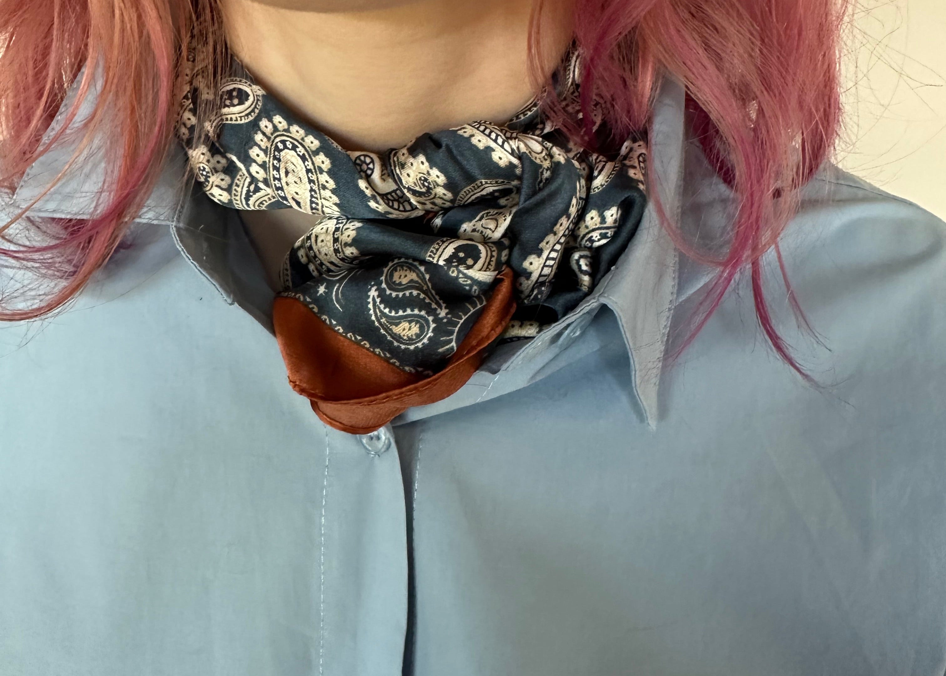 A close-up of a person wearing a dark paisley neck scarf with a brown leather-like trim, styled under a light blue button-up shirt.