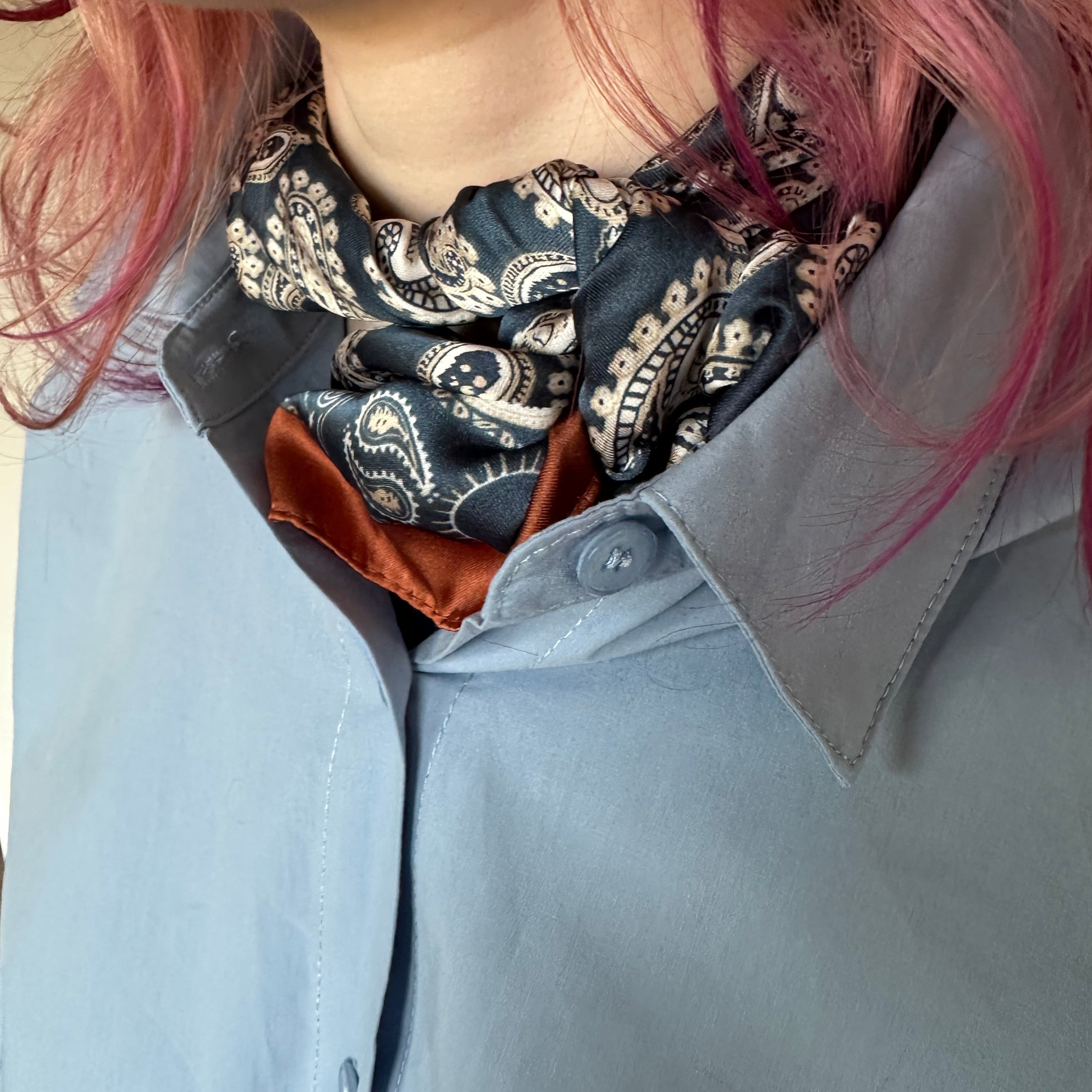 A close-up of a person wearing a light blue button-up shirt with a patterned scarf tied around the neck. The scarf has a navy, beige, and white paisley design with a rust-orange inner layer visible at the collar.