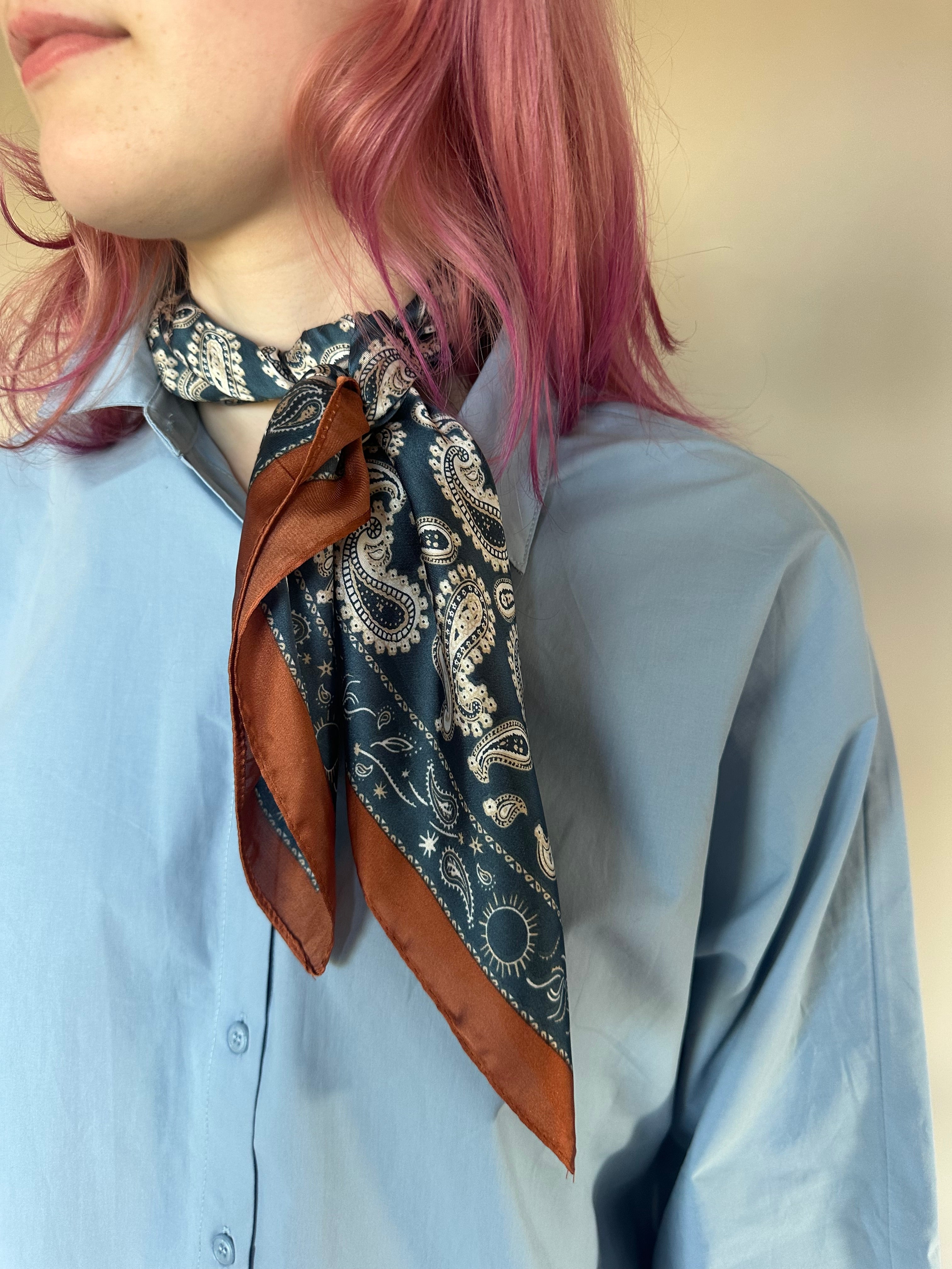 A close-up of a person wearing a light blue button-up shirt and a navy blue paisley scarf with brown borders, styled around the neck.