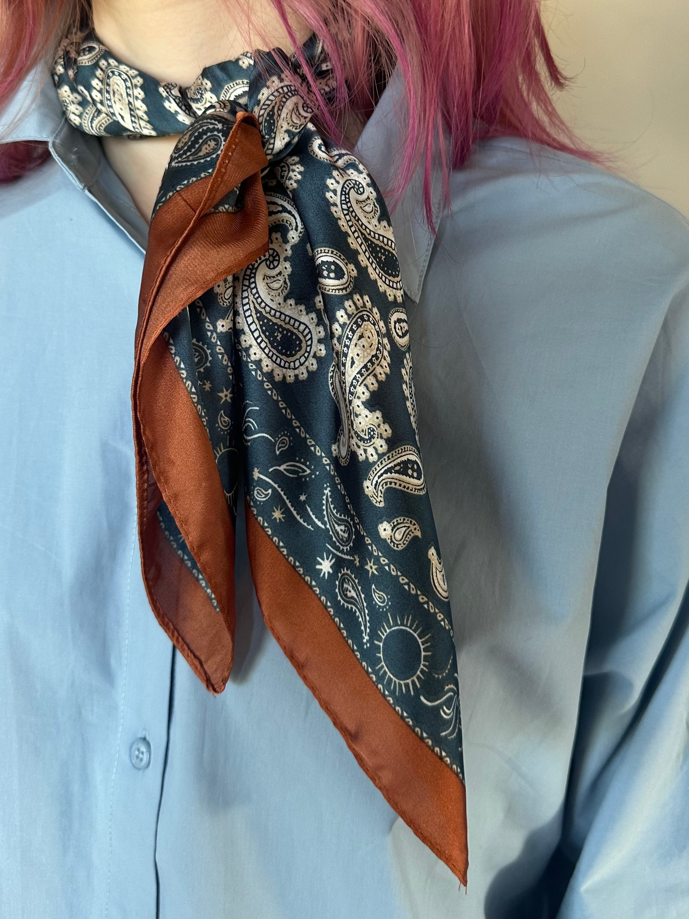 A close-up of a person wearing a light blue button-up shirt and a dark paisley neck scarf with rust-brown borders and cream detailing. The person has pink hair.