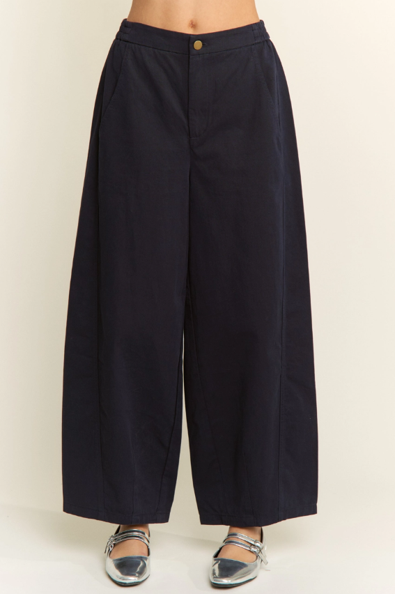 A person wearing dark navy wide-leg cropped pants with a single gold button at the waistband, paired with metallic silver flats, against a plain light beige studio background.