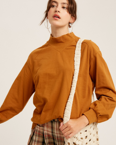 A woman wearing a mustard brown mock neck long-sleeve top, plaid pants, and carrying a chunky cream crochet shoulder bag against a light beige studio background.