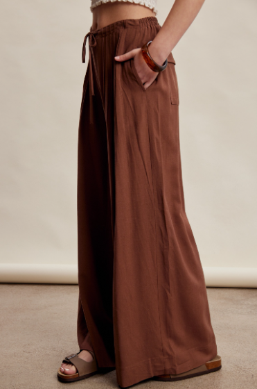 A person wearing loose, wide-leg brown pants with a drawstring waist and side pockets, paired with brown sandals, photographed against a plain beige studio backdrop.