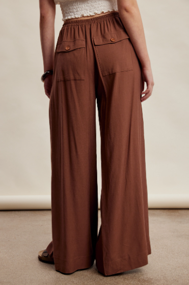 A person shown from the back wearing wide-leg brown pants with an elastic waistband, flap pockets, and a white textured top, standing against a plain beige studio backdrop.