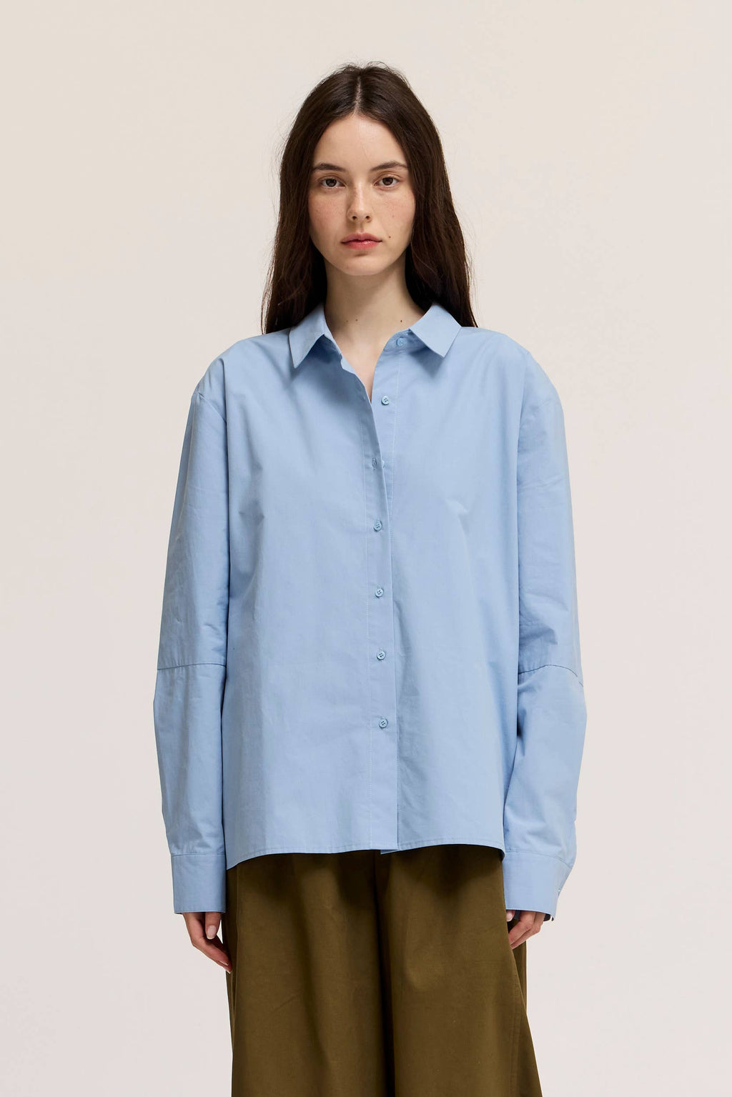 Woman wearing a light blue shirt and brown pants against a beige background